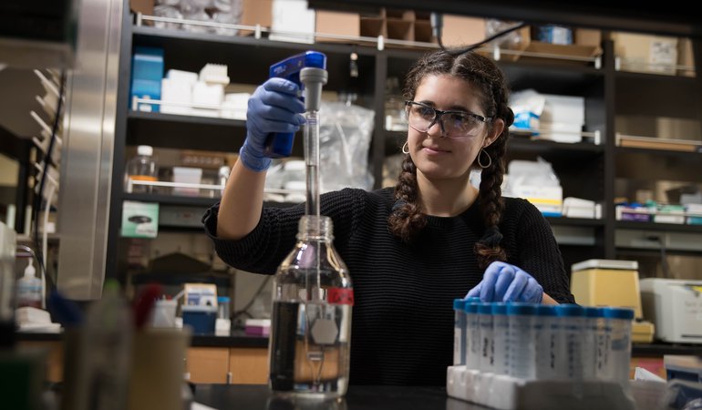 Gabby, in a lab, wearing goggles and gloves to extract liquid from a bottle.