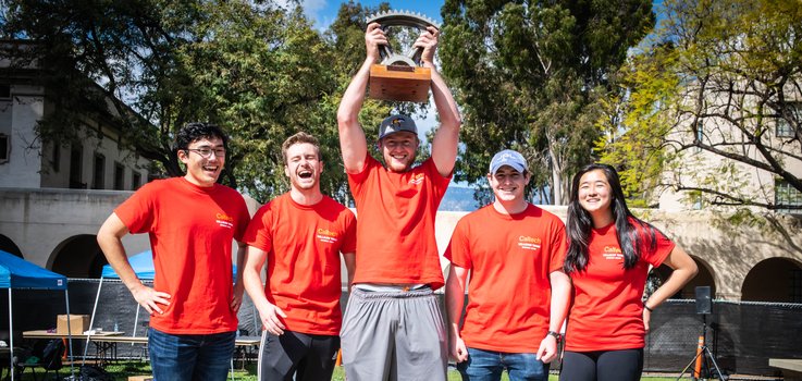 Five students stand in a line after the ME72 Competition. Student in the center is holding the trophy above their head.