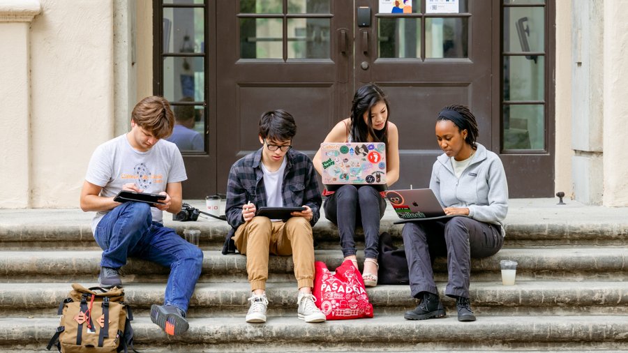 Four students sitting on stairs looking at their computers and discussing work.