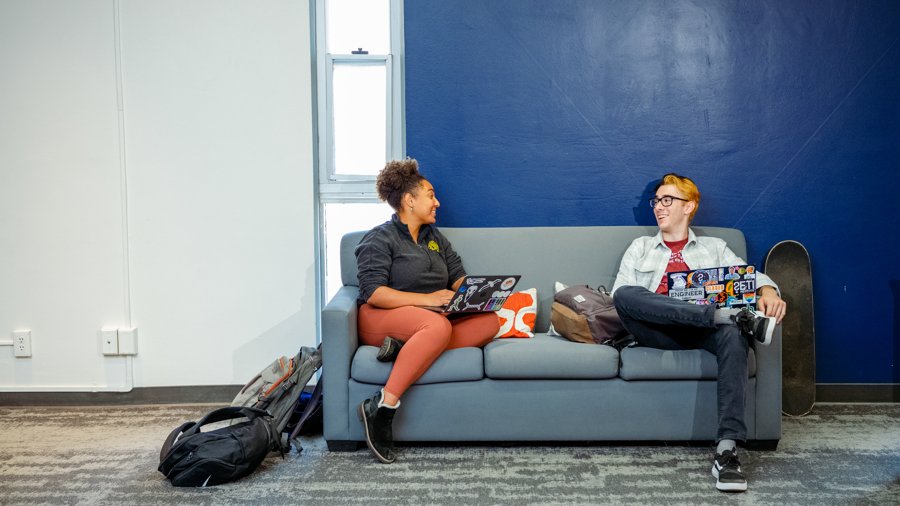 Two students sitting on a gray couch with a blue and white wall