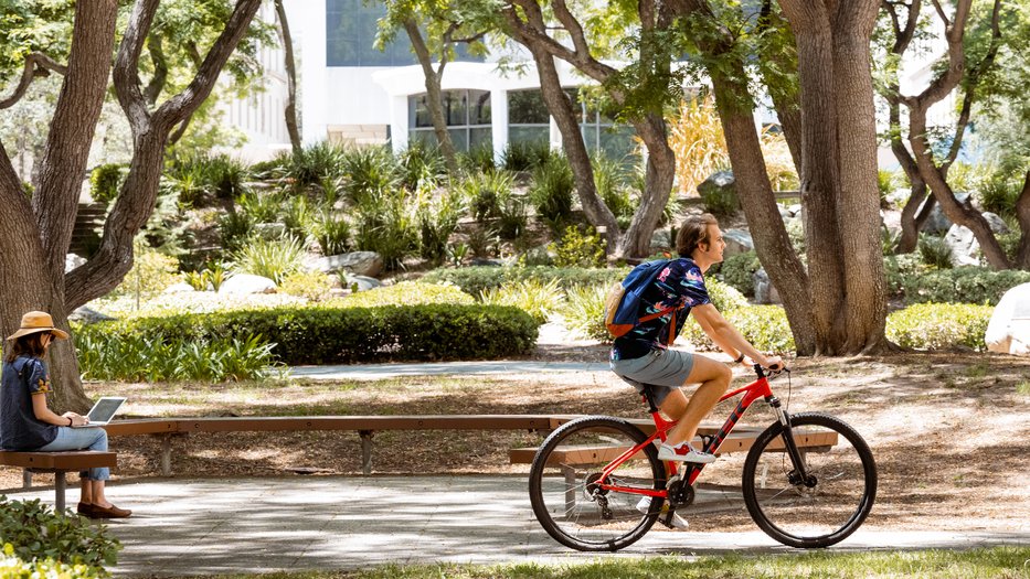 A student riding a red bicycle and a person sitting working on their laptop