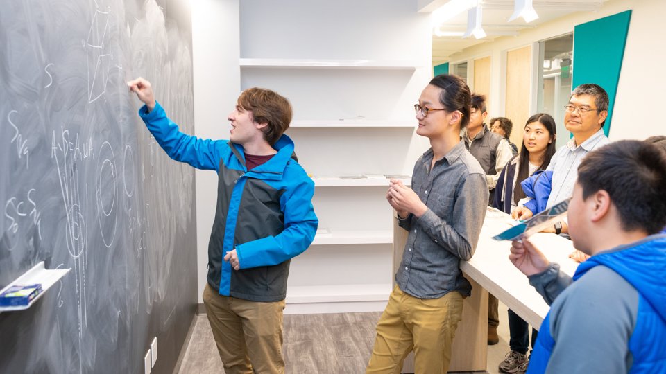 Students write on a blackboard in a hallway with people watching.