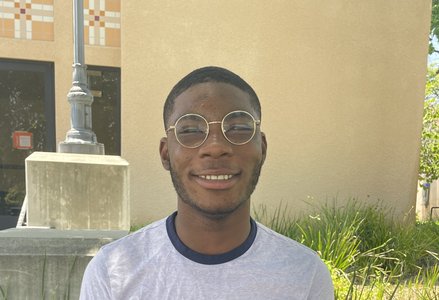 Young Black man with short hair wearing round glasses and a light grey t-shirt with a blue collar, smiling in front of a terracotta building