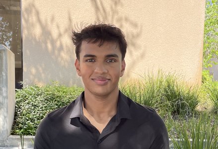 Young South Asian man with short hair and a black polo shirt smiling in front of a terracotta building