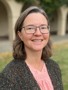 Woman with brown hair to her shoulders tucked behind her ears wearing rectangular glasses in a grey cardigan over a pink blouse