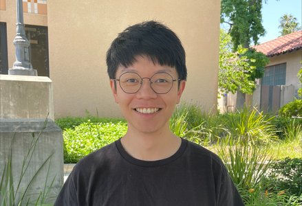Young man with short black hair wearing glasses and a black t-shirt smiling, standing in front of a terracotta colored building