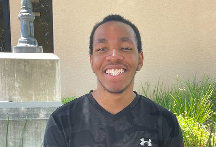 Young Black man with short hair wearing a black Under Armour smiling in front of a terracotta building