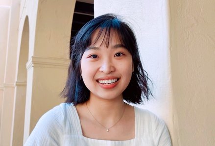 Young Asian woman with a short, black bob and bangs and a flowy square-neck top smiling in front of Caltech eaves