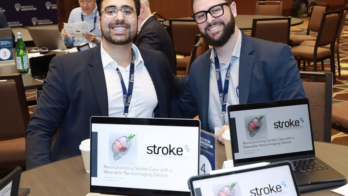 Two smartly dressed young men at a conference table, laptops open with "Stroke Dx" on them.