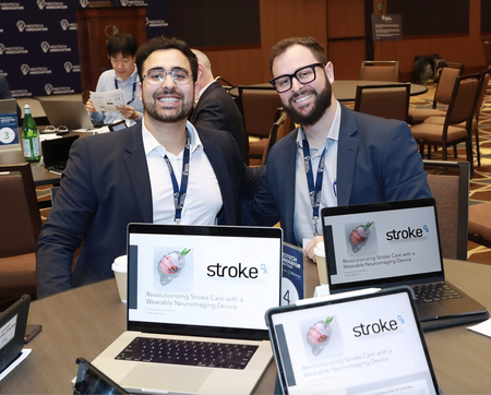 Two smartly dressed young men at a conference table, laptops open with "Stroke Dx" on them.
