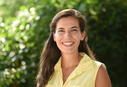 A young woman with long, medium brown hair wearing a pale yellow top smiling in front of a hedge