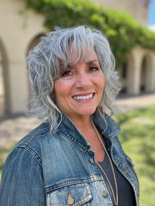 A woman with a wavy grey angled bob, wearing a jean jacket, smiling