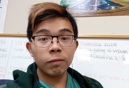 Young man with short, dark brown hair with a highlight at the front and glasses wearing a light green shirt and a dark green hoodie in front of a whiteboard and a poster of fireworks at Caltech