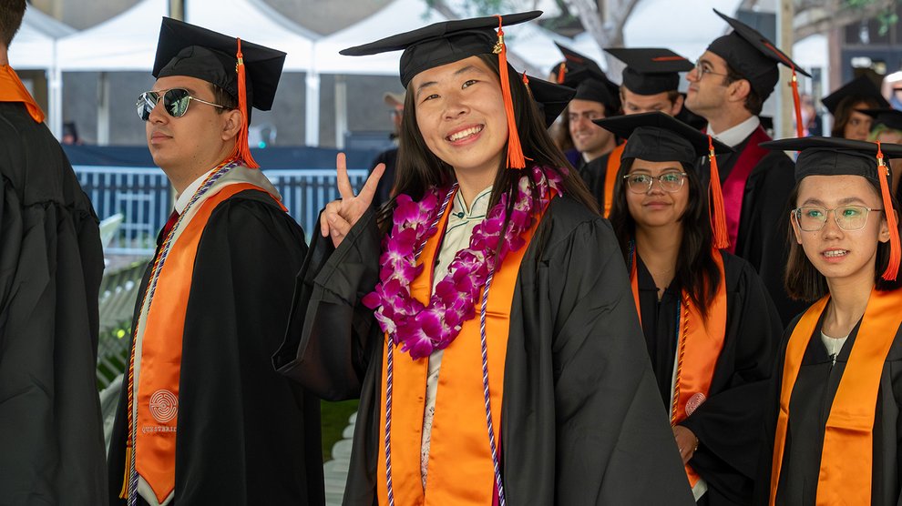 A group of graduates in caps and gowns processes into their commencement ceremony. One woman smiles at the camera and flashes the peace sign.