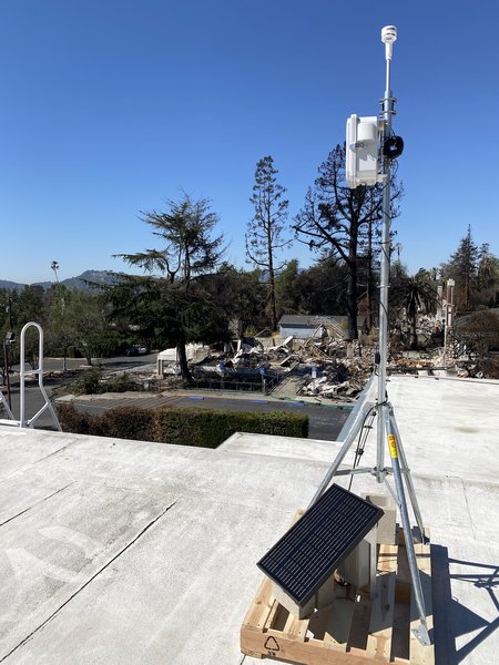 A QuantAQ sensor installed on a rooftop. Burned property from the fire is clearly visible in the background.