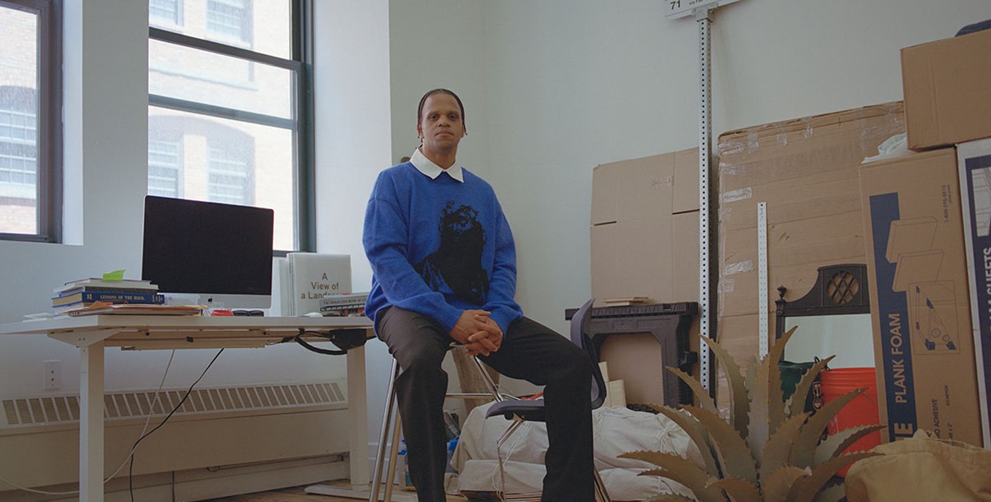 A man in a blue sweater, black pants, and black shoes sits with his hands in his lap looking into the camera. He is seated on a stool in an art studio, surrounded by cardboard, elements of a sculpture, and a desk with a computer and books.