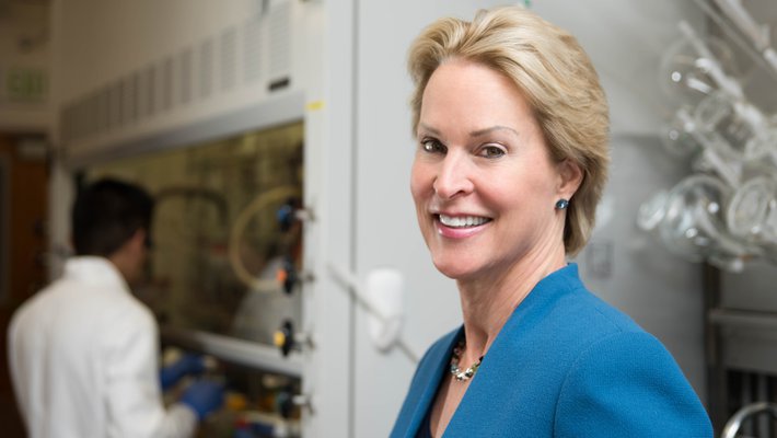 A portrait of Frances Arnold in her laboratory. She wears a blue blazer and smiles at the camera. In the background are laboratory glassware and a researcher at work.