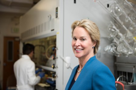 A portrait of Frances Arnold in her laboratory. She wears a blue blazer and smiles at the camera. In the background are laboratory glassware and a researcher at work.