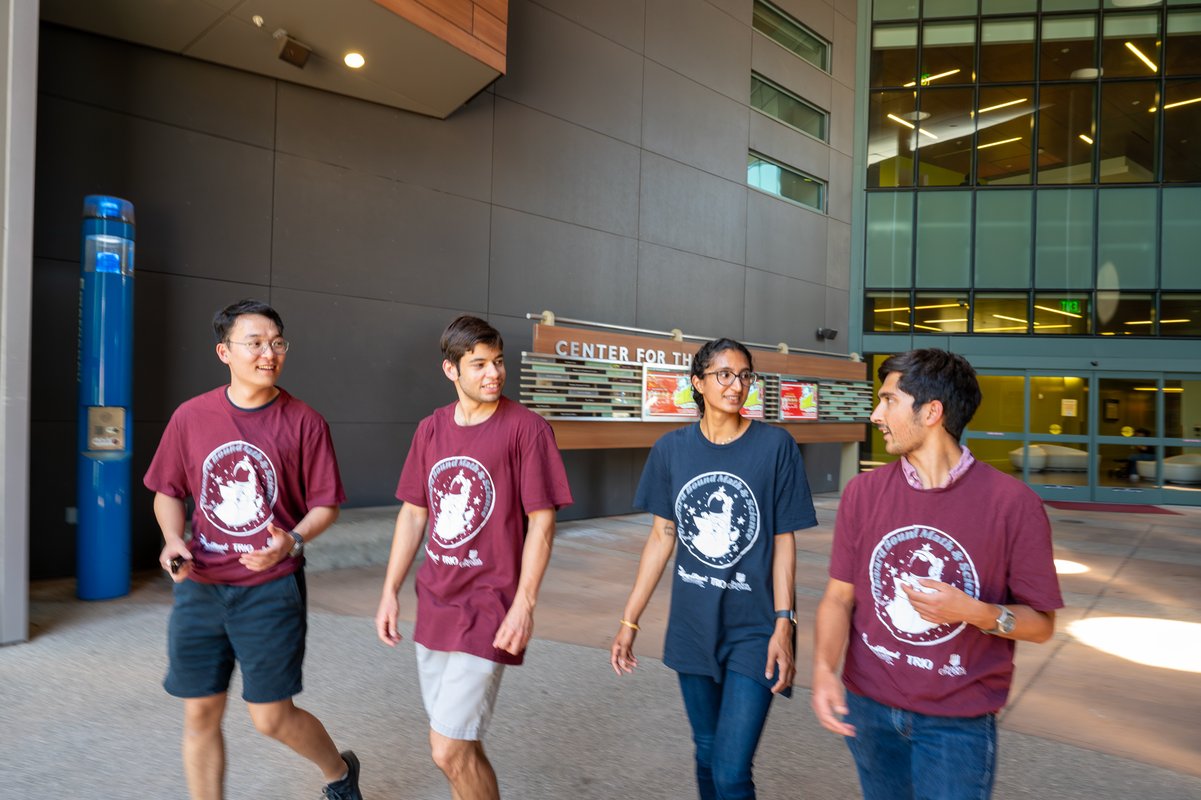 Four people walk toward the camera with a modern building behind them.