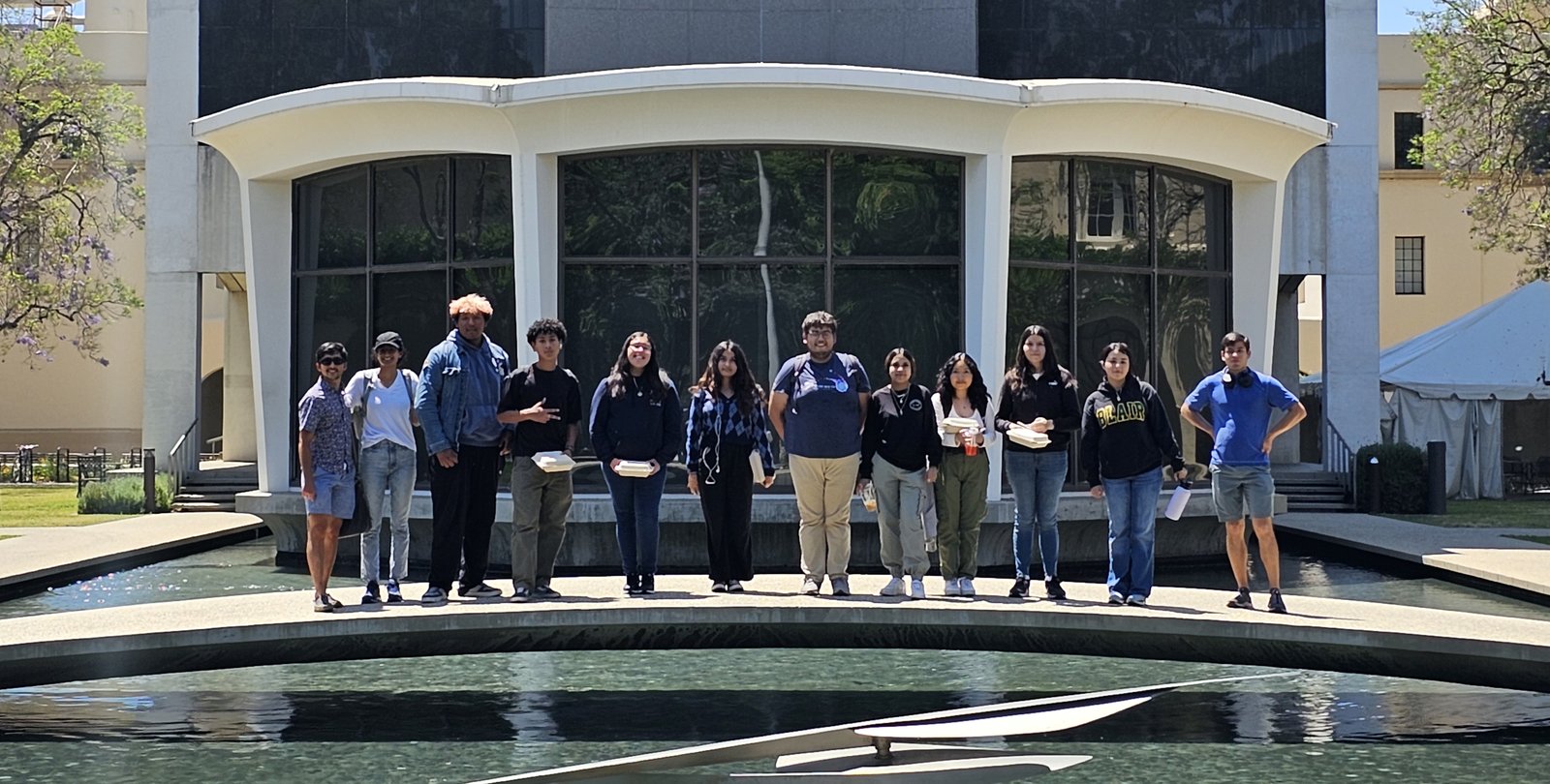 Twelve people stand on a bridge over a reflecting pool in front of a midcentury building.