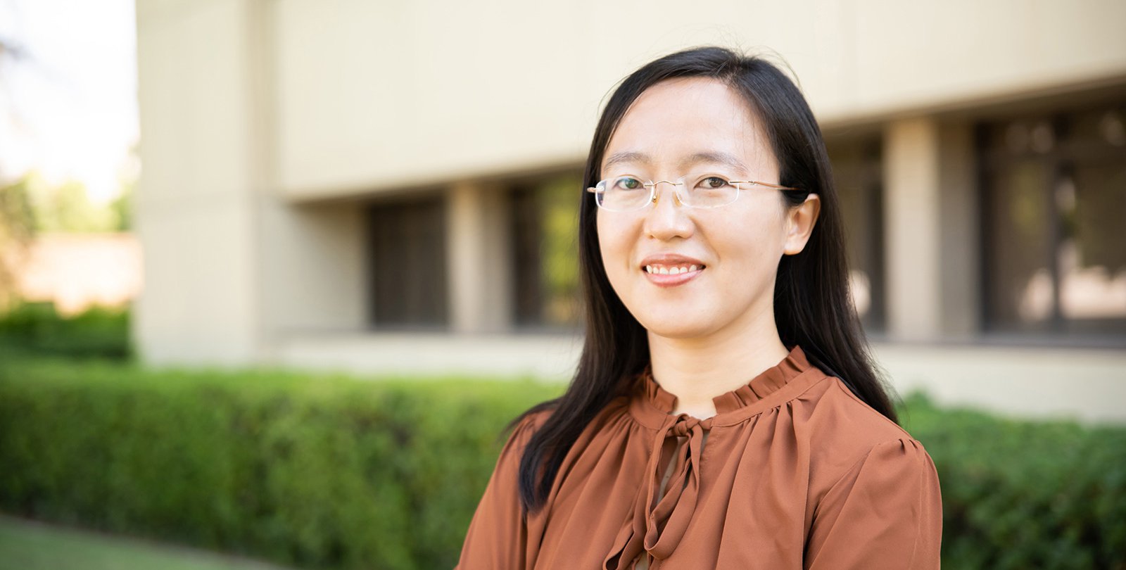 A portrait of Shasha Chong. She smiles at the camera and stands in front of a campus building with trees and grass in the background.