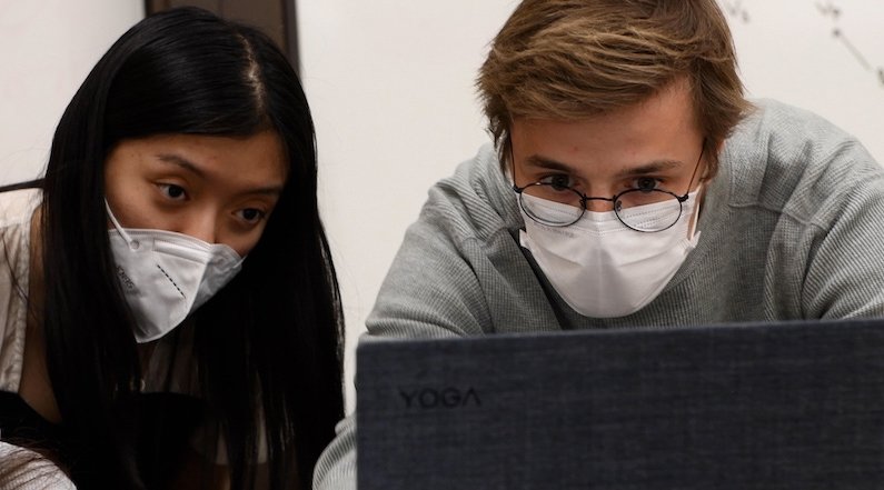 Two people wearing KN95 masks lean in to a laptop as one types, with a whiteboard in the background.