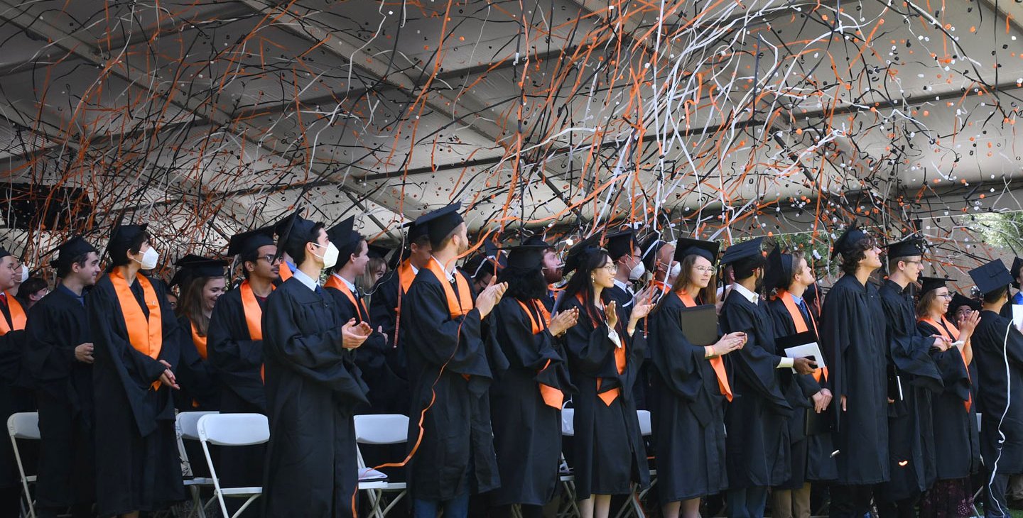 Students in caps and gowns celebrate commencement as orange confetti flies in the air.