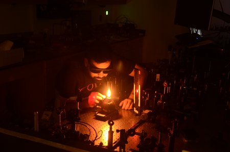 A man with a beard holds a device while looking into a flame within an optical instrument setup in a lab.