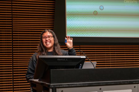 A person at a podium smiles as she holds a small vial full of blue powder.