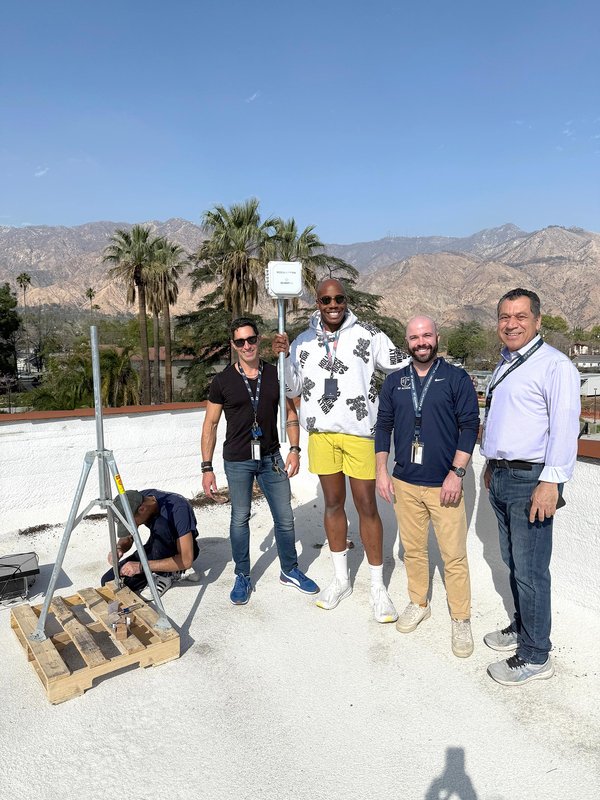 Four men stand close to a QuantAQ sensor on a rooftop as another man sitting nearby works to install the sensor. Palm trees and the San Gabriel Mountains are in the background.