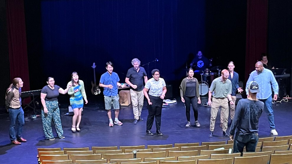 A group of about 10 people perform on a stage and Brian Brophy stands in the audience and directs them