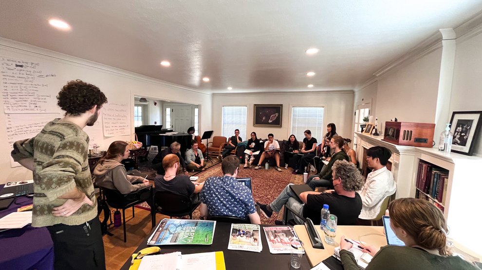A group of about 20 people sit in chairs in a circle inside a room in a house.