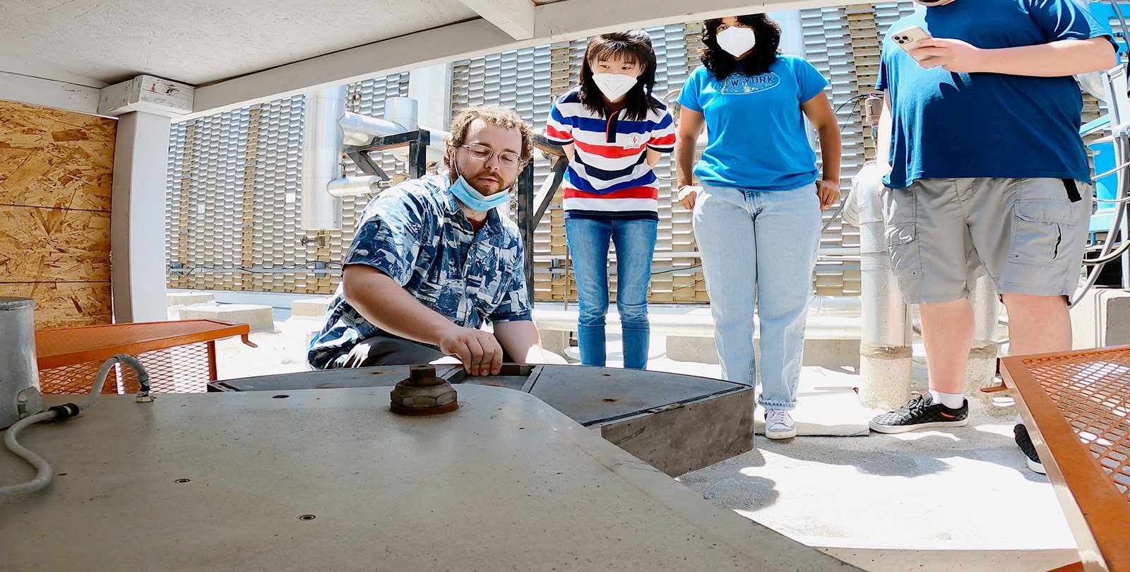 Four people are on the flat, white asphalt roof of a building. One squats in the foreground touching a large metal wedge as three others look on, two watching intently and the third taking photographs with a phone.