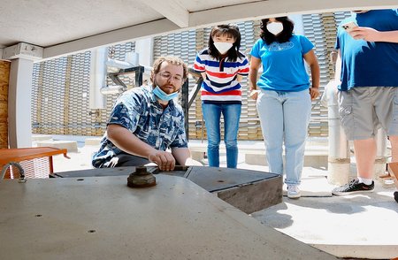 three people watch as one kneels and touches a large wedge-shaped object