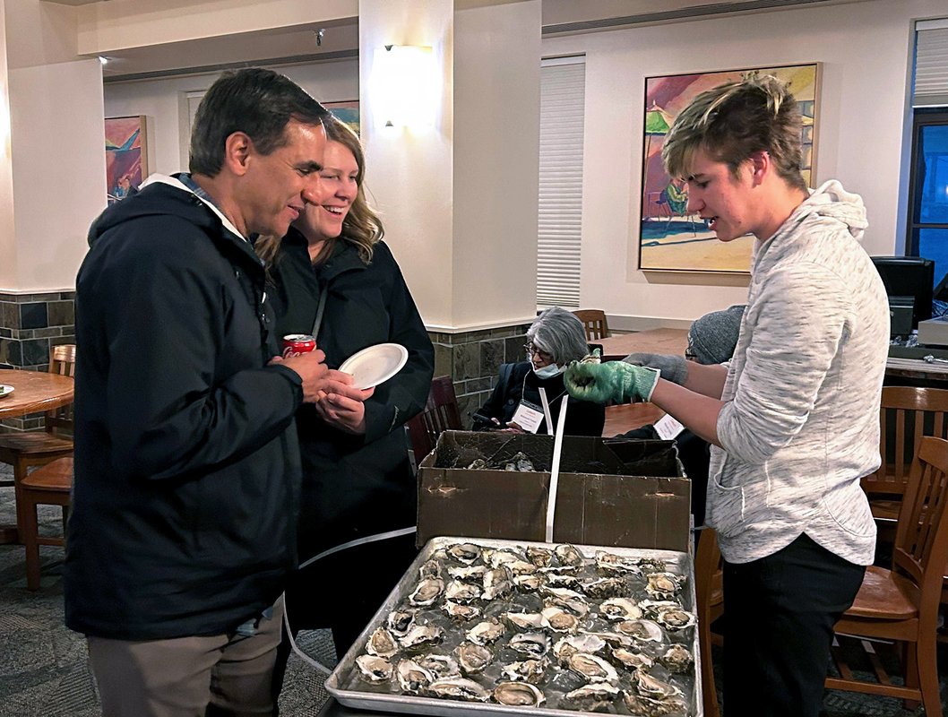 A student demonstrates how to shuck an oyster to two parents.