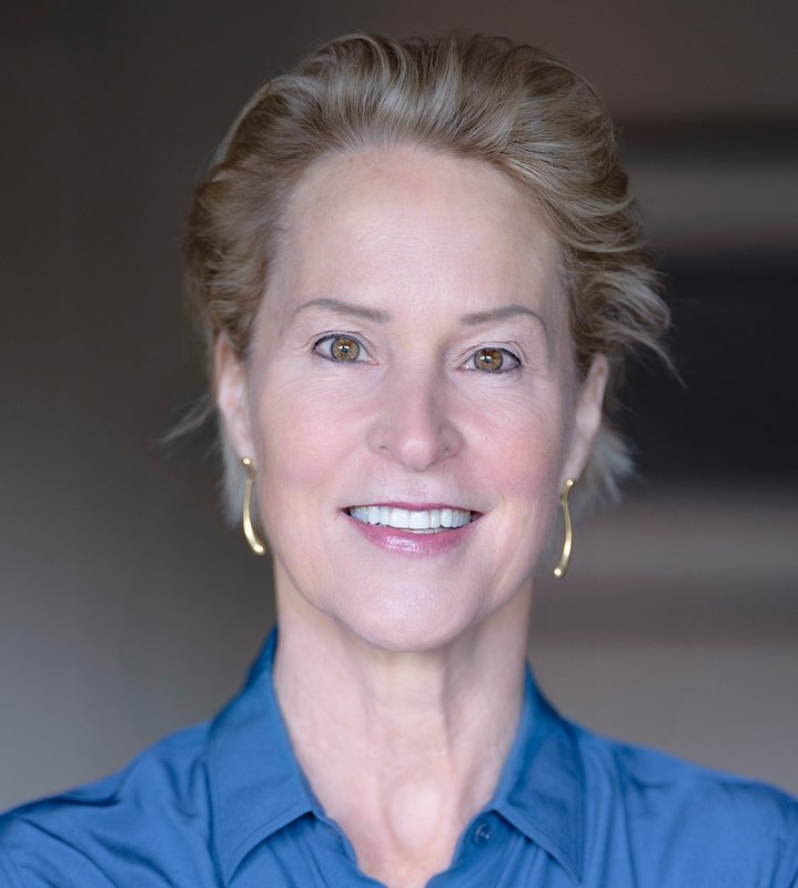 Head shot of a woman with short blond hair wearing a blue collared shirt and gold earrings.