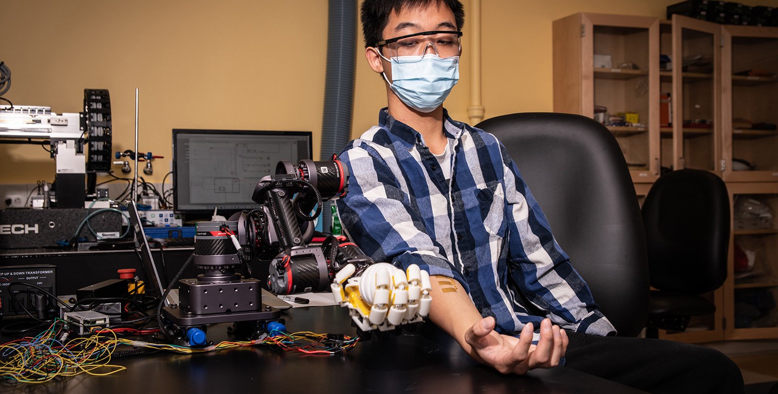 A human sits at a table flexing his hand. Sensors are attached to the skin of his forearm. A robotic hand next to him mimics his motion.