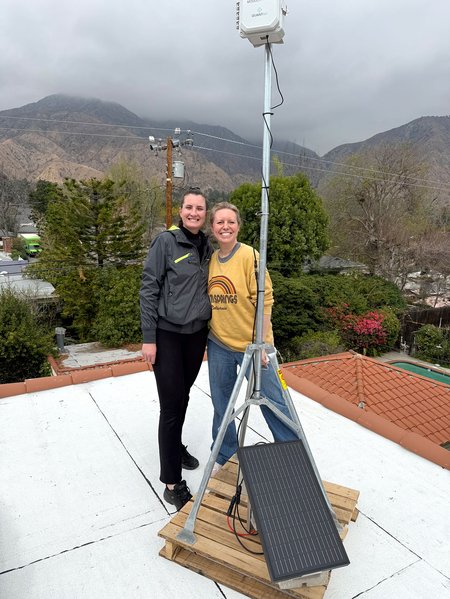Two women stand behind a particulate air quality sensor mounted like a tripod on a roof in front of stormy looking mountains.