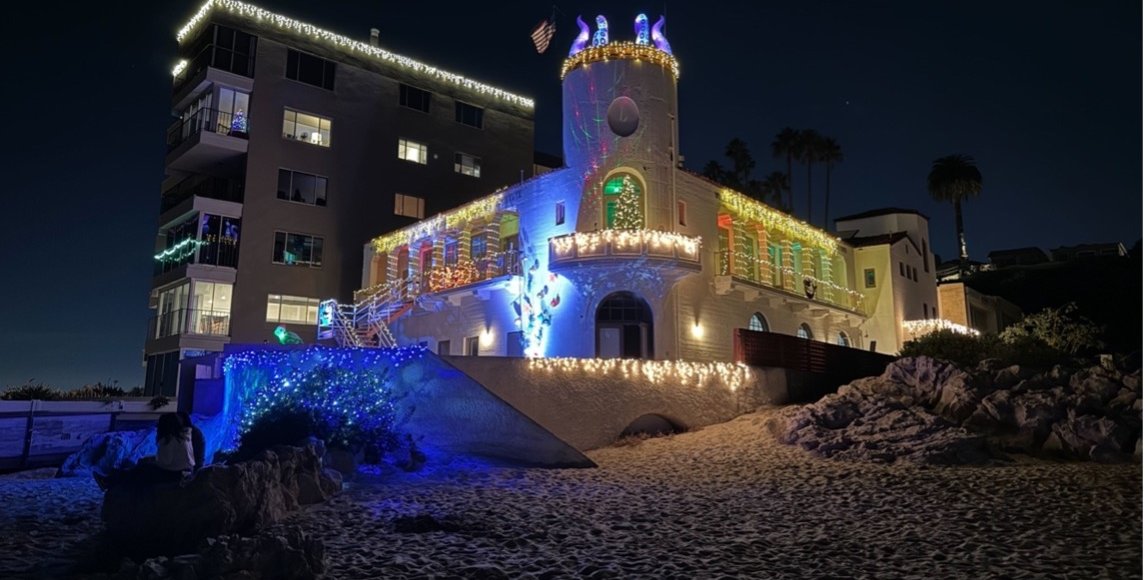 A large stucco building decorated with holiday lights stands above a beach, in front of decorated condominiums