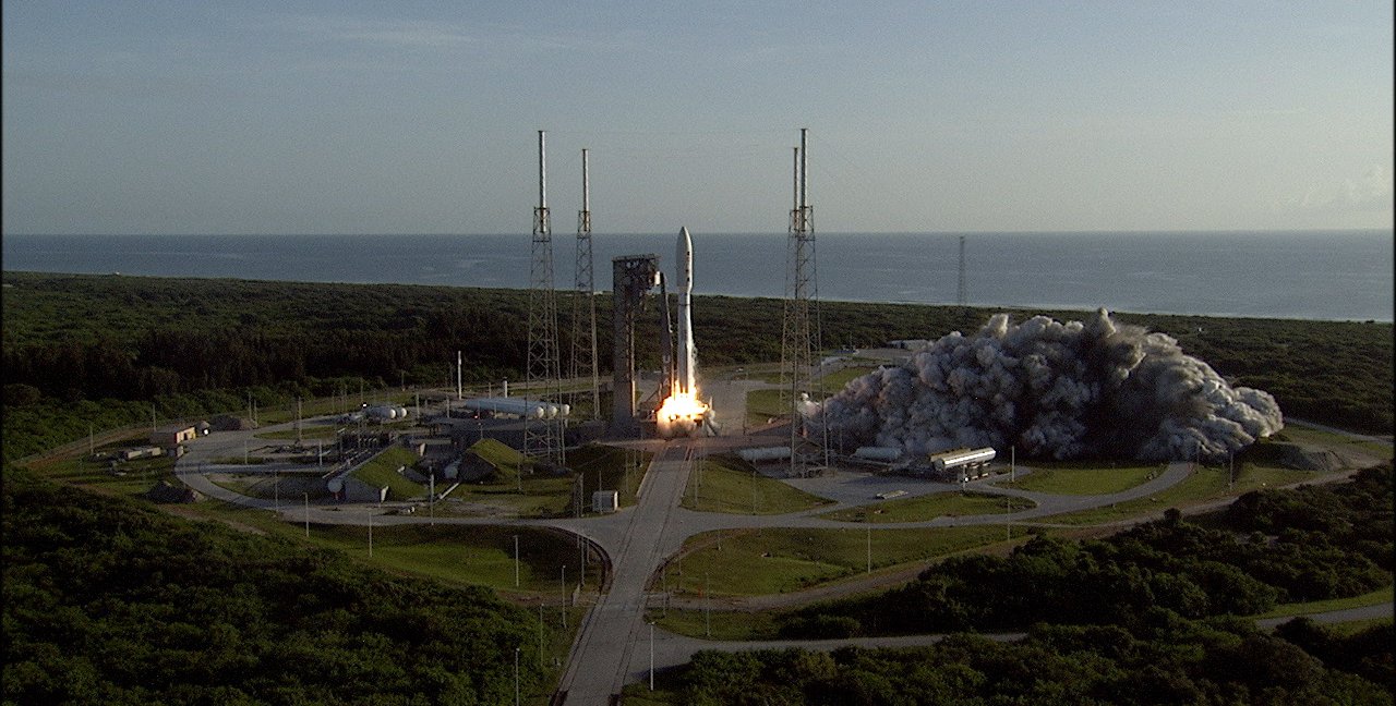 A rocket begins launching from its pad, sending smoke and flames outward. Ocean appears in the background.