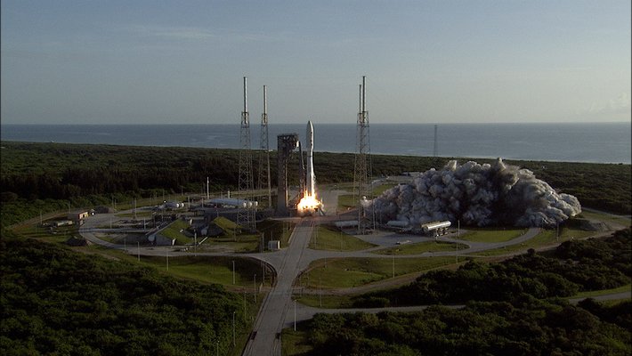 A rocket begins launching from its pad, sending smoke and flames outward. Ocean appears in the background.