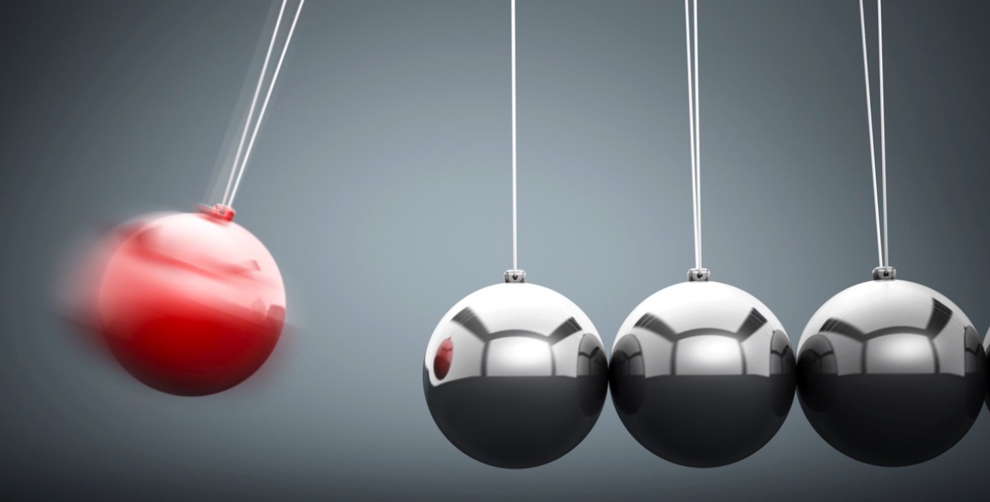 A Newton's cradle toy is shown with the far left, red ball in movement, headed toward hitting the next silver ball.