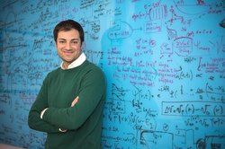 A portrait of Marco Bernardi. He stands in front of a blue chalkboard covered in writing with his arms crossed.