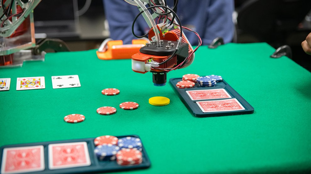 A robotic arm plays a game of blackjack. Red playing cards and red/white and blue/white poker chips as well as a solid yellow chip are on the green felt table.