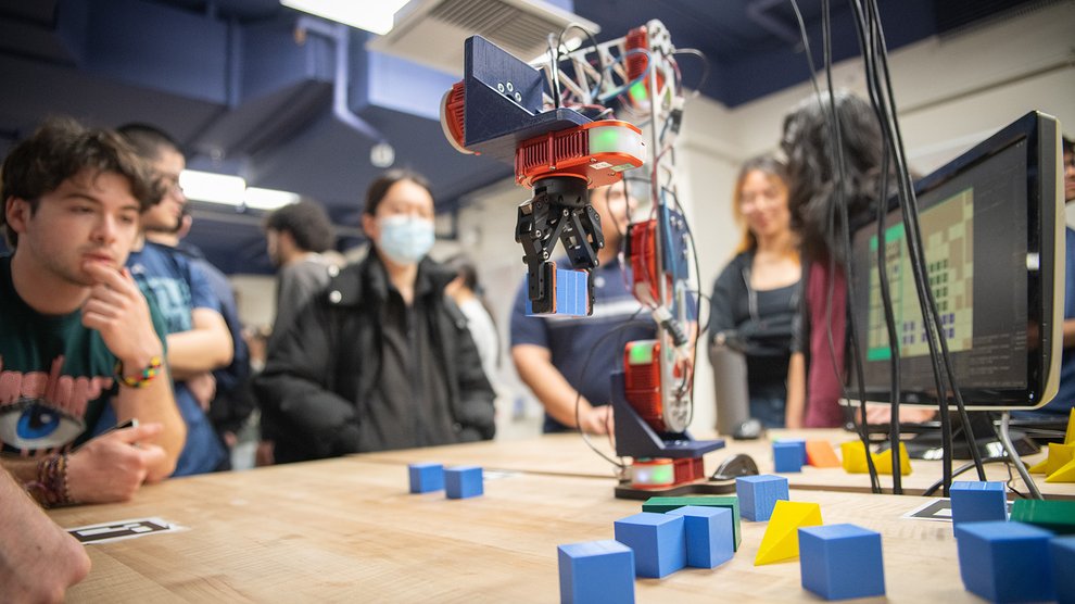 A robotic arm is pinching and lifting a blue block off the tabletop.