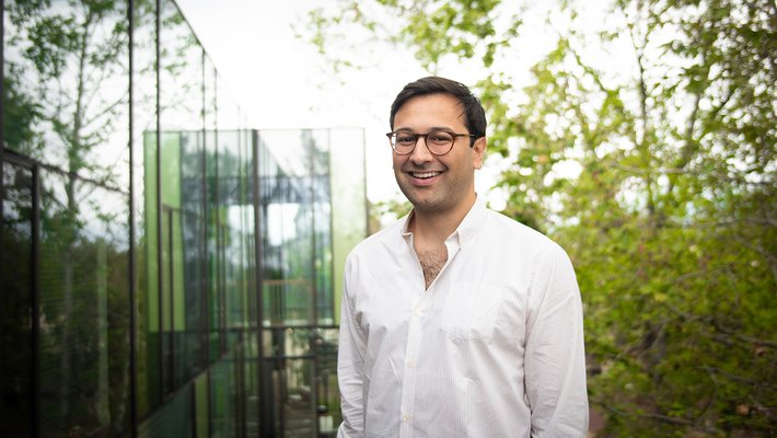 A portrait of Eric Mazumdar. He is outdoors with his hands in his pockets and smiles at the camera.