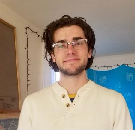 A portrait of Noah Dayton Peake. He is indoors and smiling at the camera. He wears glasses and has a short beard.