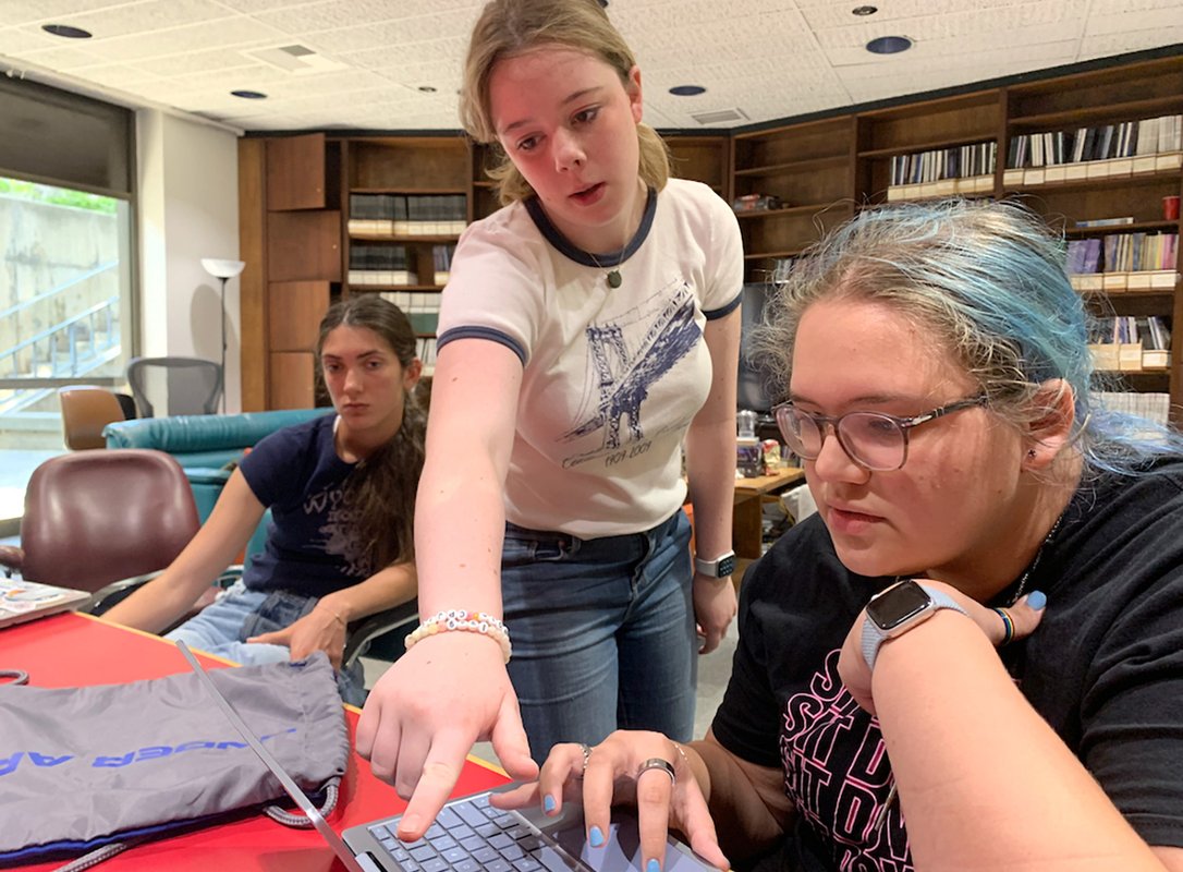 Three high school students sti at a table in a library. Two are discussing something on a computer screen.