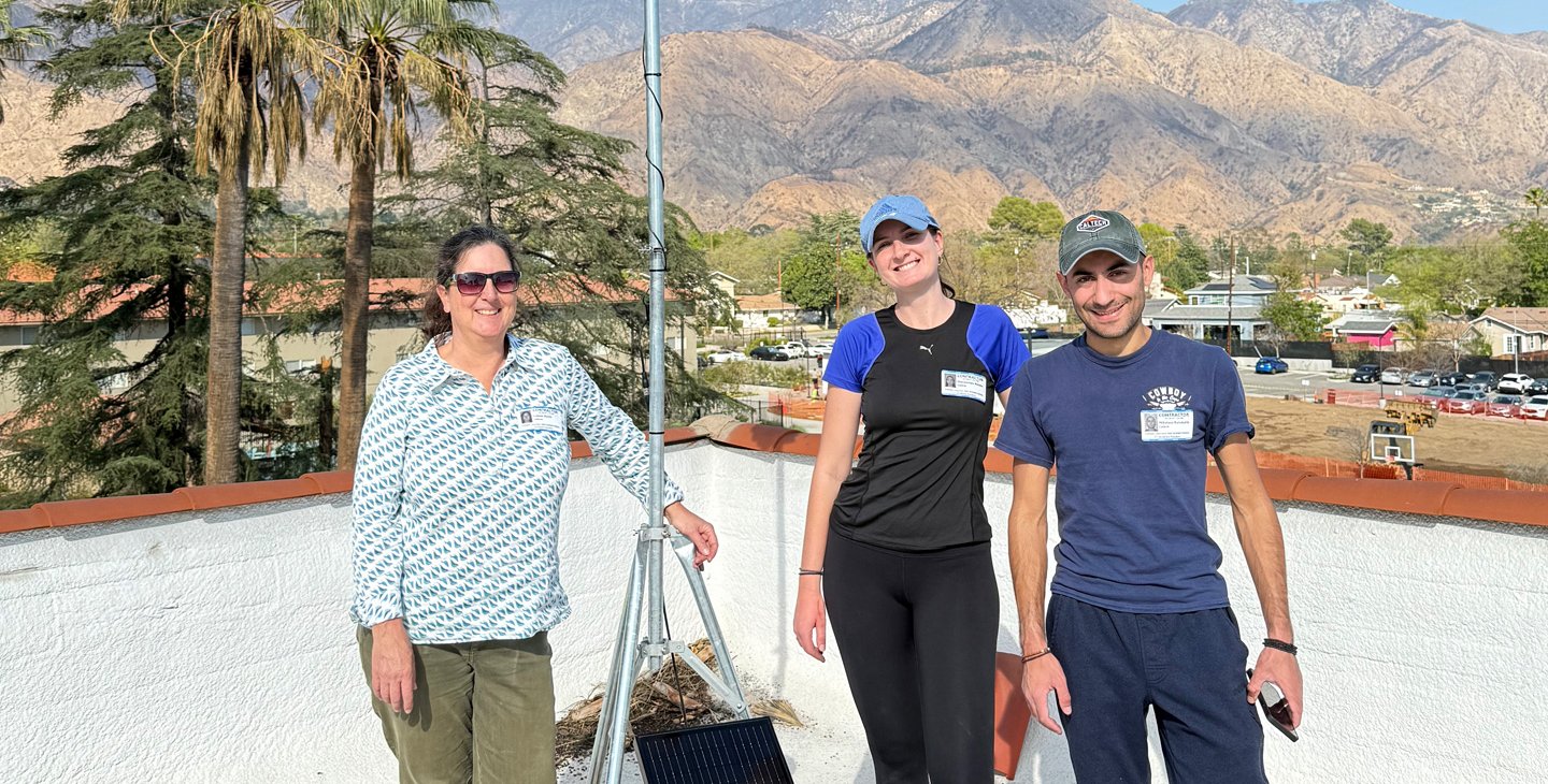 Two women and a man pose with an installed PHOENIX sensor on a rooftop with the San Gabriel Mountains in the background.