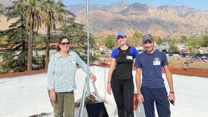 Two women and a man pose with an installed PHOENIX sensor on a rooftop with the San Gabriel Mountains in the background.